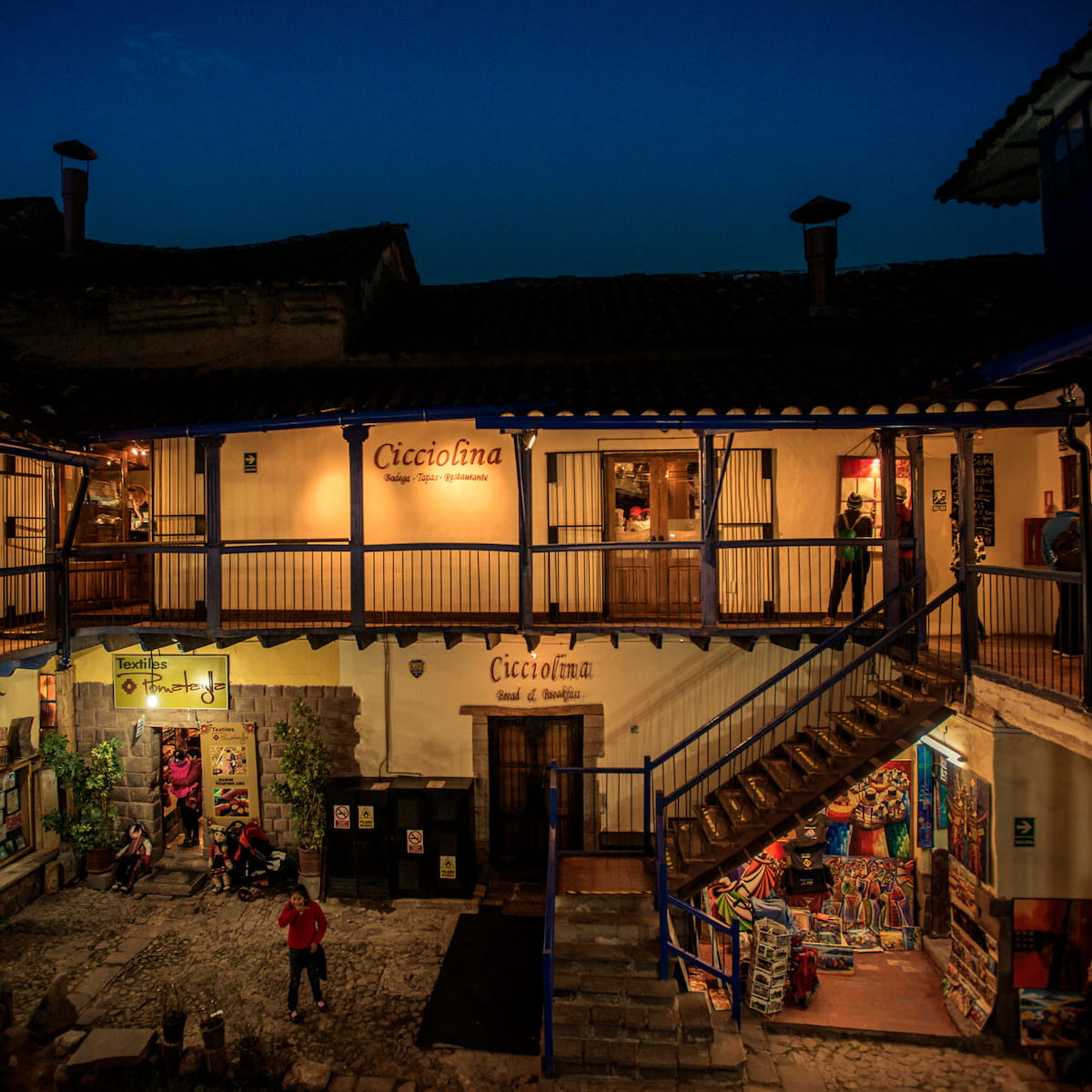 Exterior view of Cicciolina restaurant in Cusco during the evening with ambient lighting, featuring a balcony, signage, and local textiles shop below - The Cusco Guide